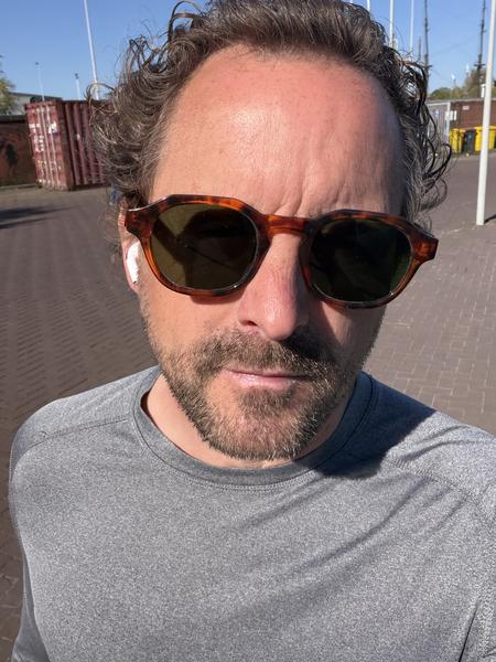 A man takes a close-up selfie outdoors on a bright, sunny day, wearing sunglasses and a grey long-sleeved shirt